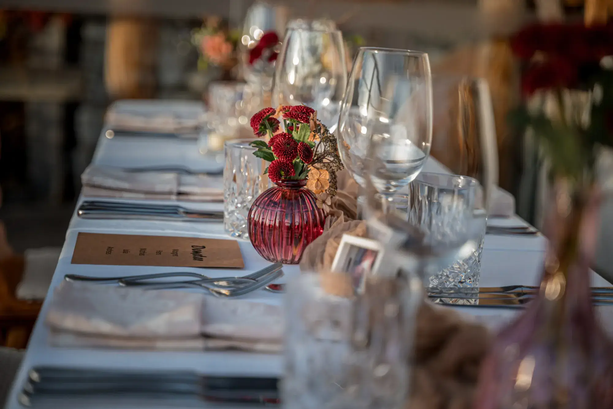 Festtafel-Tischdeko mit roten Blumen in rubinroter Vase und Menükarte im warmen Abendlicht der Stöttlalm